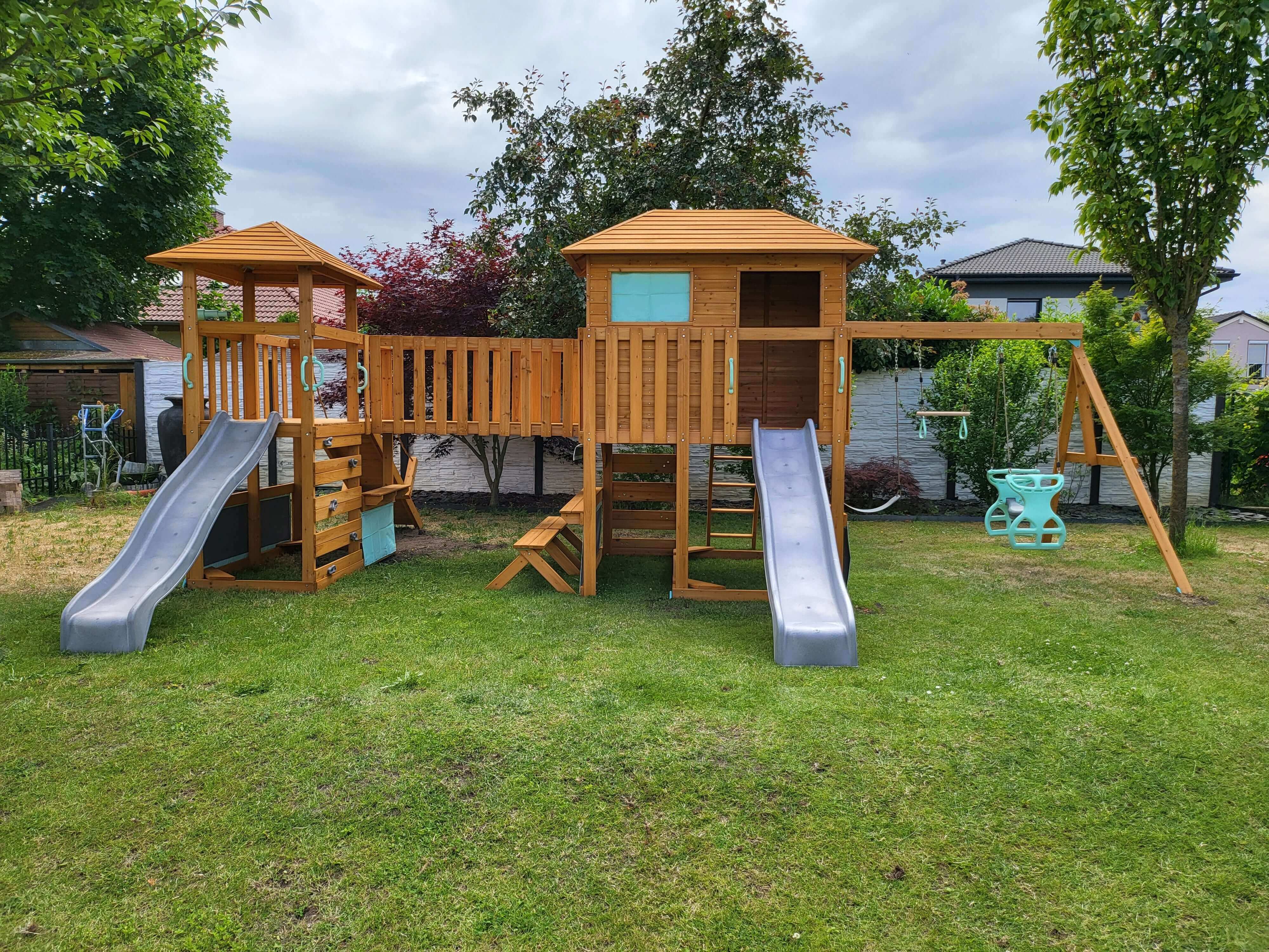 A Petit Seal Adventure Paradise Whale Play Tower - Exciting Playland Experience is nestled in a grassy backyard, sparking children's creativity. The setup features two slides, a central playhouse with railings, a small ladder, and swings. Surrounded by trees, garden furniture, and a shed under cloudy skies, it's perfect for endless adventures.