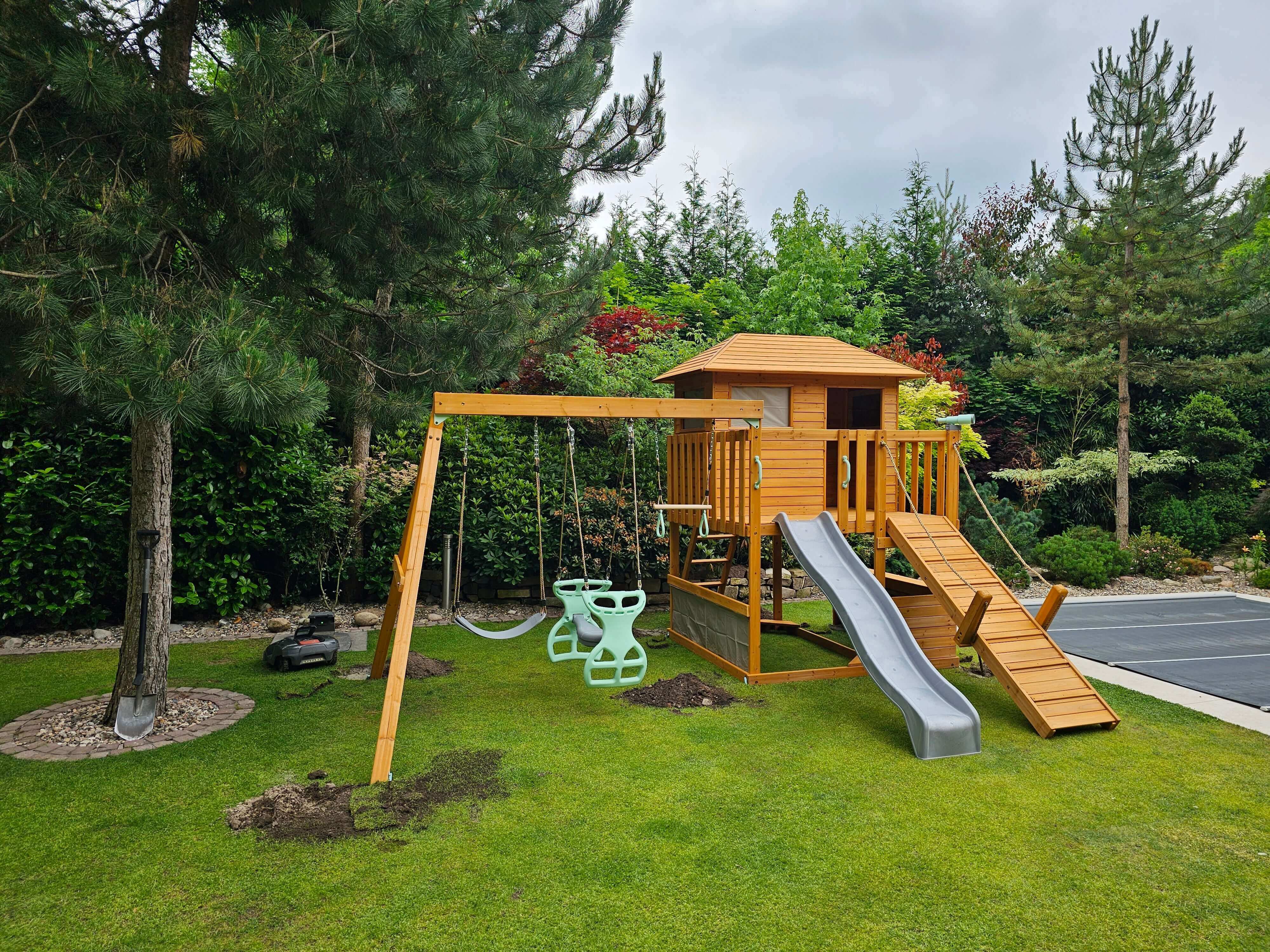 A wooden playground set in a lush green backyard featuring a treehouse with a ramp, slide, and swingset with dual green toddler swings. The surrounding area is landscaped with various trees and bushes, and a lawn mower sits nearby. The sky is overcast with a hint of blue—a perfect spot for children's playtime adventures with the Enchanted Garden Play Tower: Unlock a Realm of Outdoor Fun and Learning with Llamazing Adventure Honey by Petit Seal.