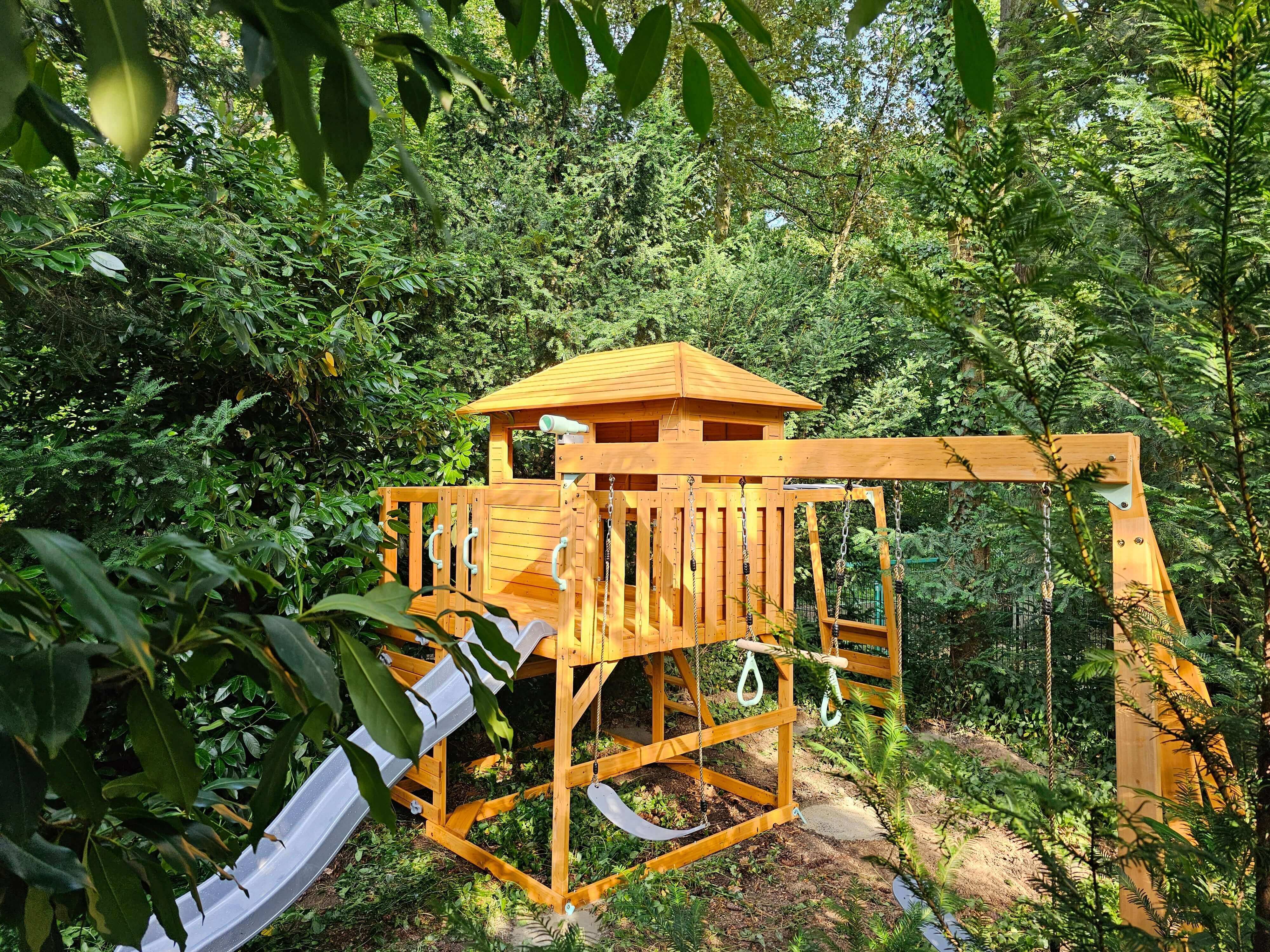 A wooden treehouse playset, known as the Hippo Hopper Play Tower: Transform Your Backyard into a Magical Playground by Petit Seal, stands amidst lush greenery. It features a slide on the left, a central playhouse structure with a roof and railing, and a swing set with climbing rope and rings on the right. This versatile play tower encourages imaginative play in a secluded, nature-rich environment.