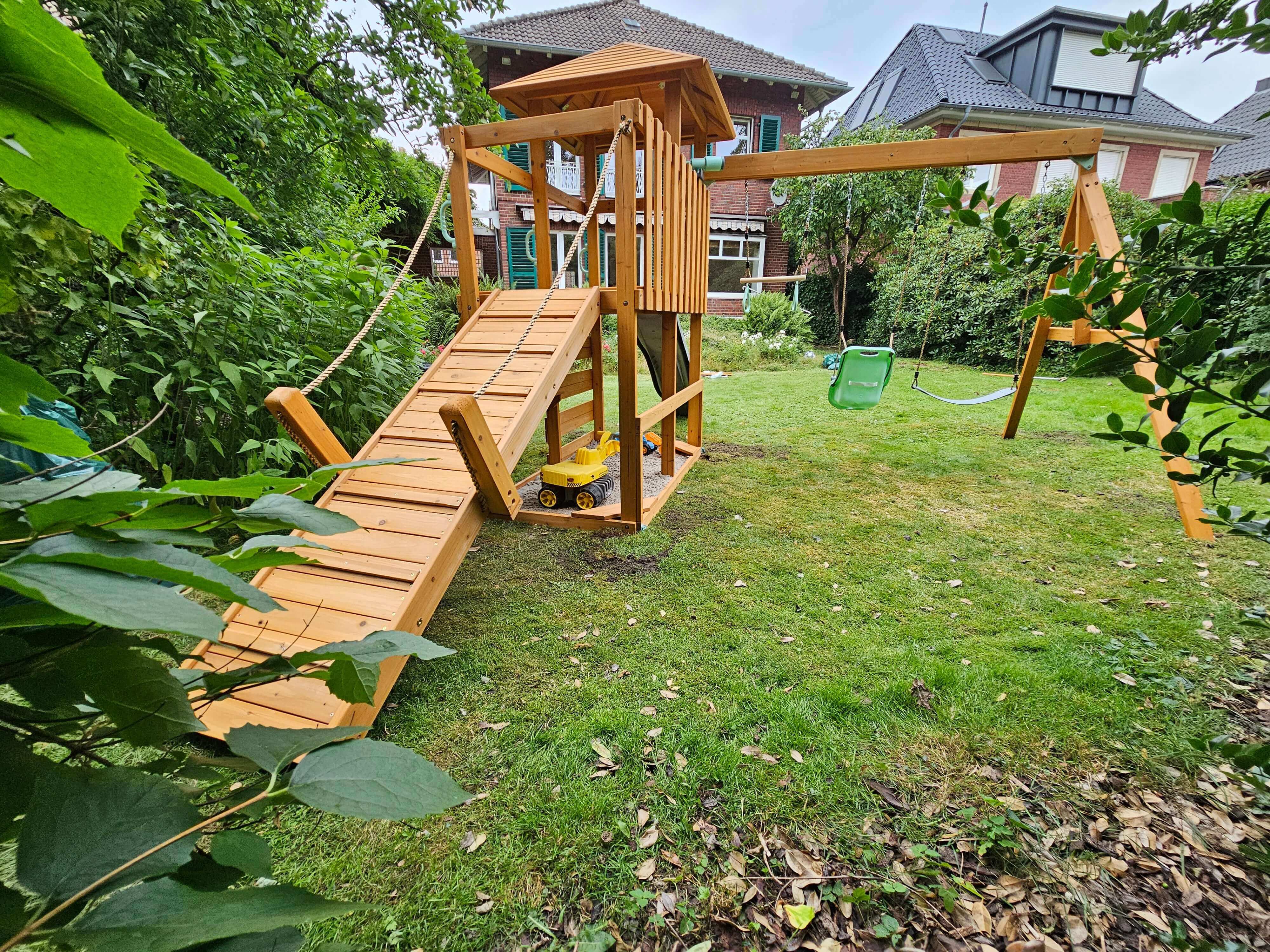 A Petit Seal Elephantastic Play Tower Honey: Infinite Fun Playground in a backyard features a climbing ramp with rope handrails leading to a raised platform, a slide on the right, and a green swing set. A yellow toy truck is on the ramp. Surrounding the playset is green grass, trees, and bushes, with houses in the background.