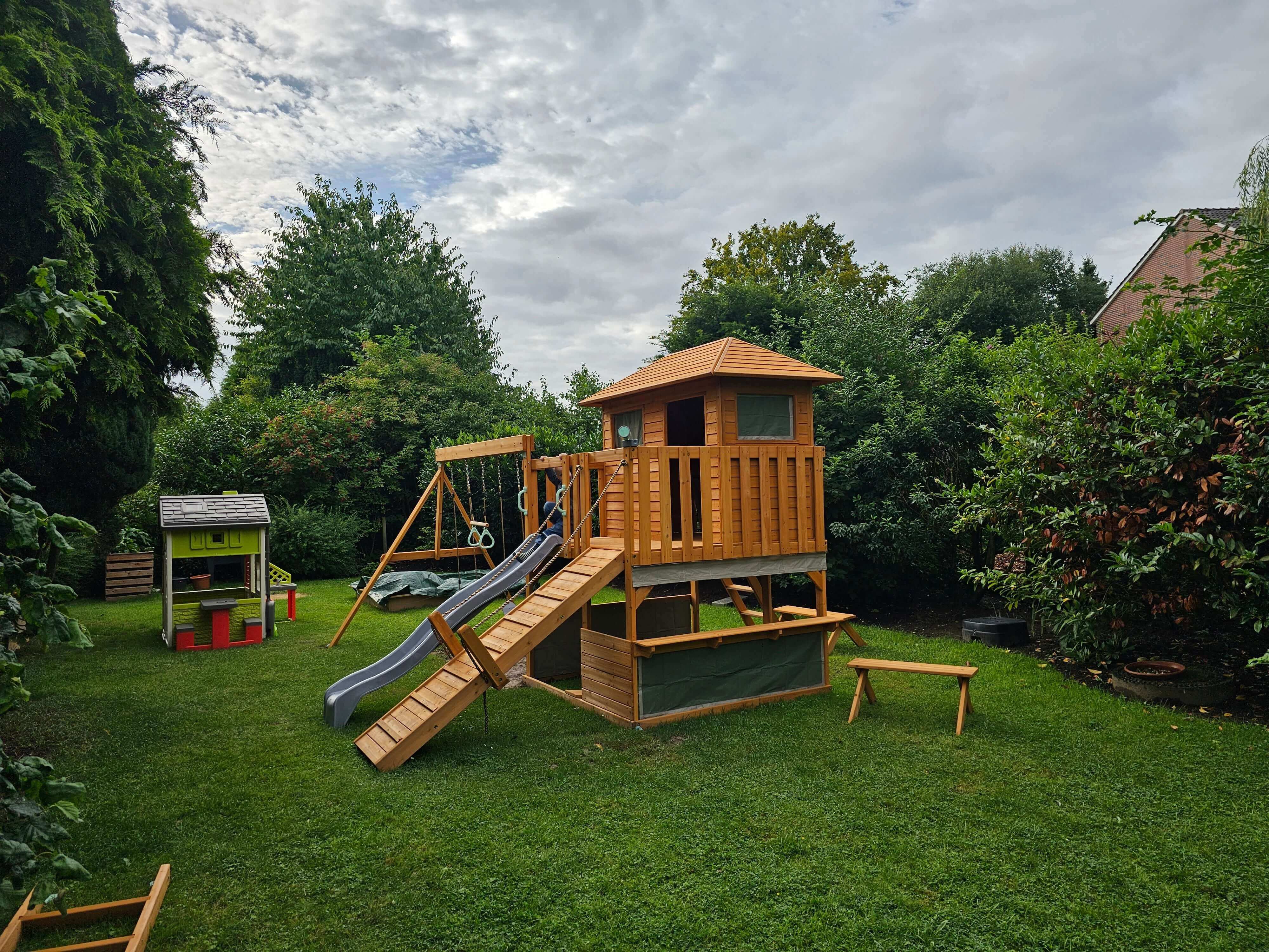 A backyard features a Enchanted Garden Play Tower: Unlock a Realm of Outdoor Fun and Learning with Llamazing Adventure Honey by Petit Seal with a climbing wall, slide, and a small house with a balcony. Nearby, there is a small plastic playhouse. Bushes, trees, and cloudy skies frame the scene. A bench sits on the green grass in the foreground to the right.