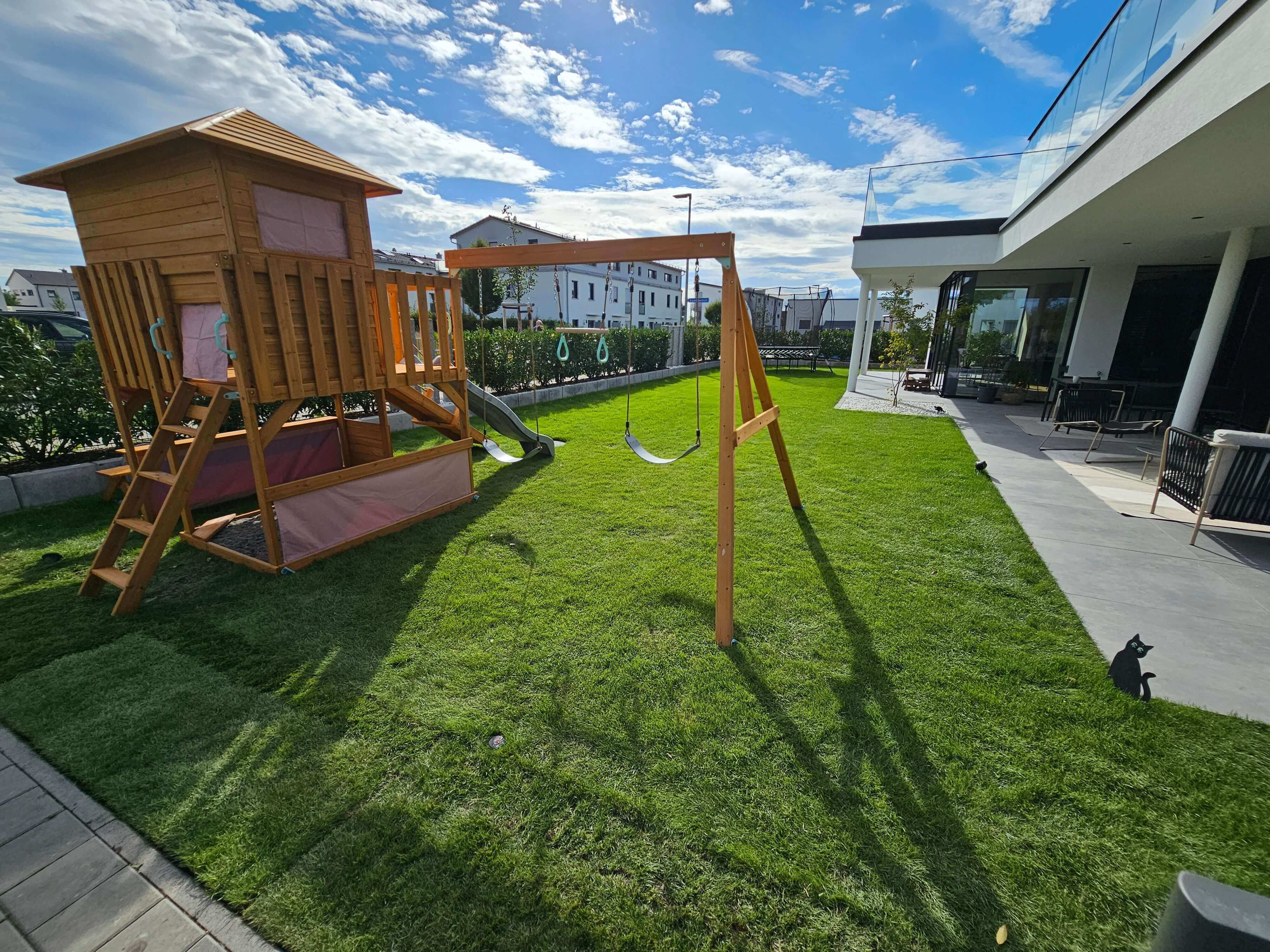 A backyard with a Petit Seal Enchanted Garden Play Tower: Unlock a Realm of Outdoor Fun and Learning with Llamazing Adventure Honey featuring a slide and swings stands on a well-manicured lawn. To the right, an outdoor patio is furnished with seating and shaded by a white modern house. The sky is partly cloudy, and a black cat sculpture can be seen on the ground by the patio.