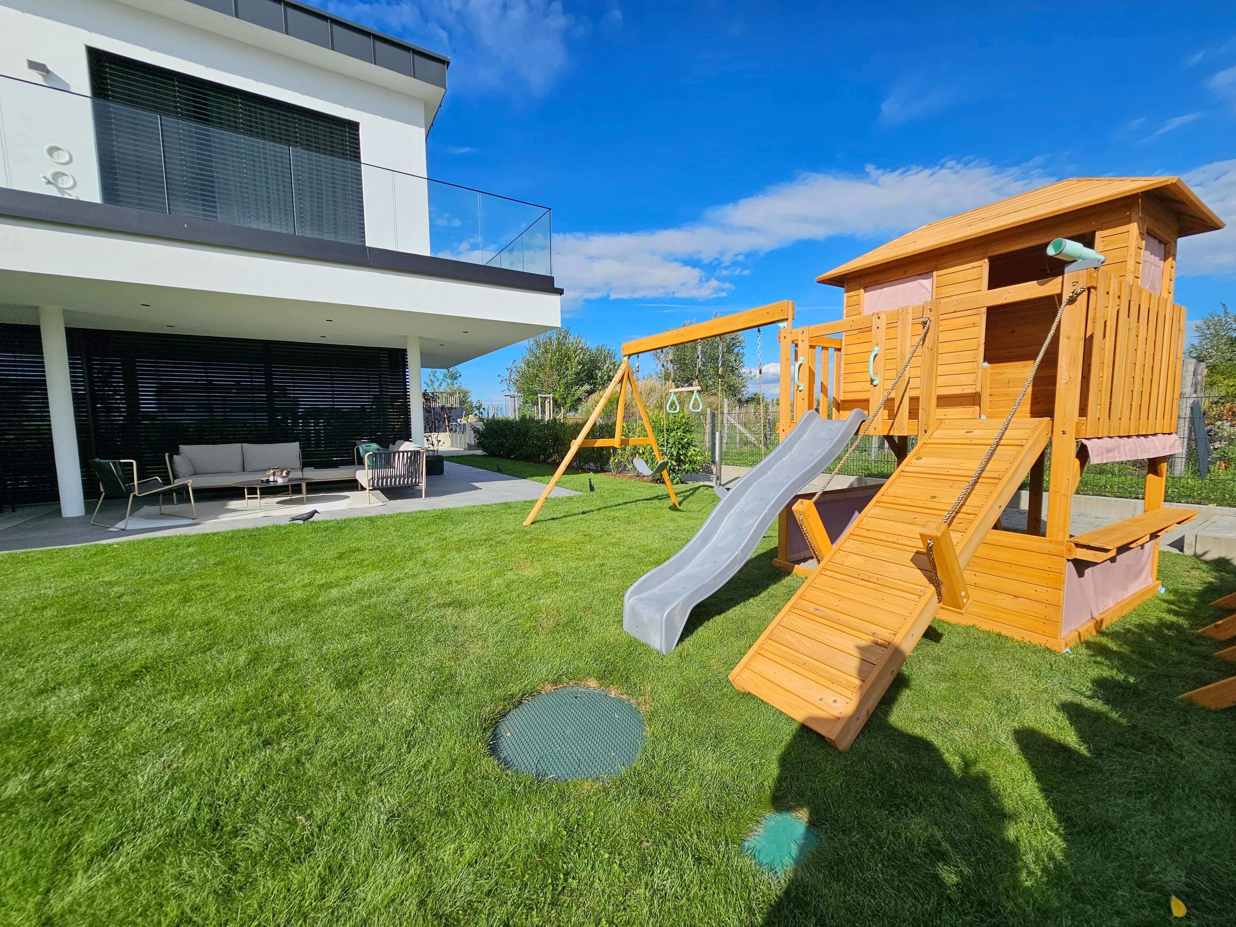 A modern backyard features an Enchanted Garden Play Tower: Unlock a Realm of Outdoor Fun and Learning with Llamazing Adventure Honey by Petit Seal with a slide, swings, monkey bars, and a climbing ramp. The lush green lawn is well-maintained. A spacious patio attached to a contemporary white house has outdoor seating, including a sofa and chairs. Clear blue sky and some clouds overhead.