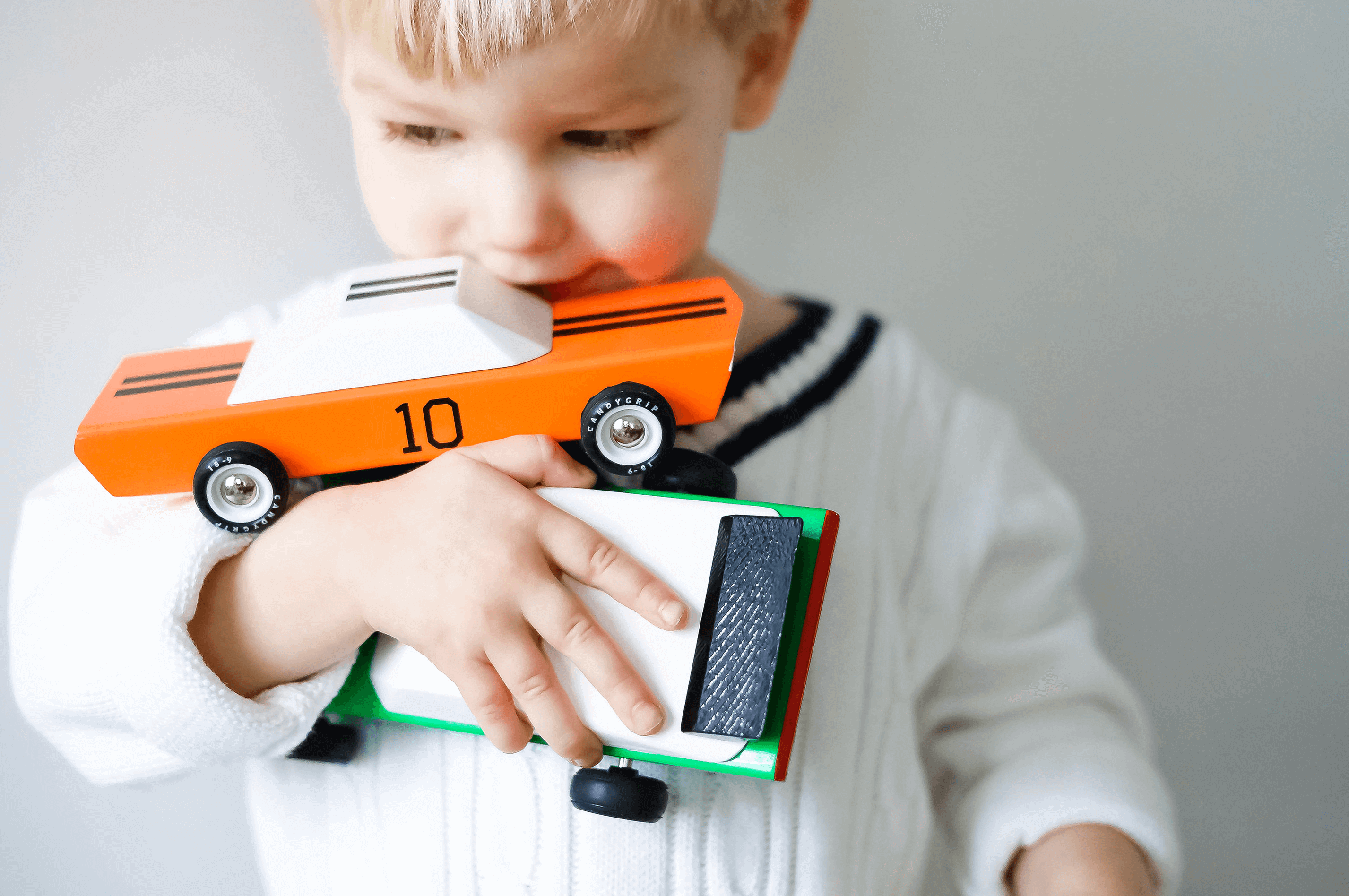 A young child with blond hair is holding two toy cars against their chest. One car, with a race-inspired design, is the American Muscle GT-10 Classic Sports Car Figurine by AMERICANA by CANDYLAB, which is orange with the number 10, and the other is white and green. The child is wearing a white sweater with dark trim and has a thoughtful expression, looking away from the camera. The background is plain and light.