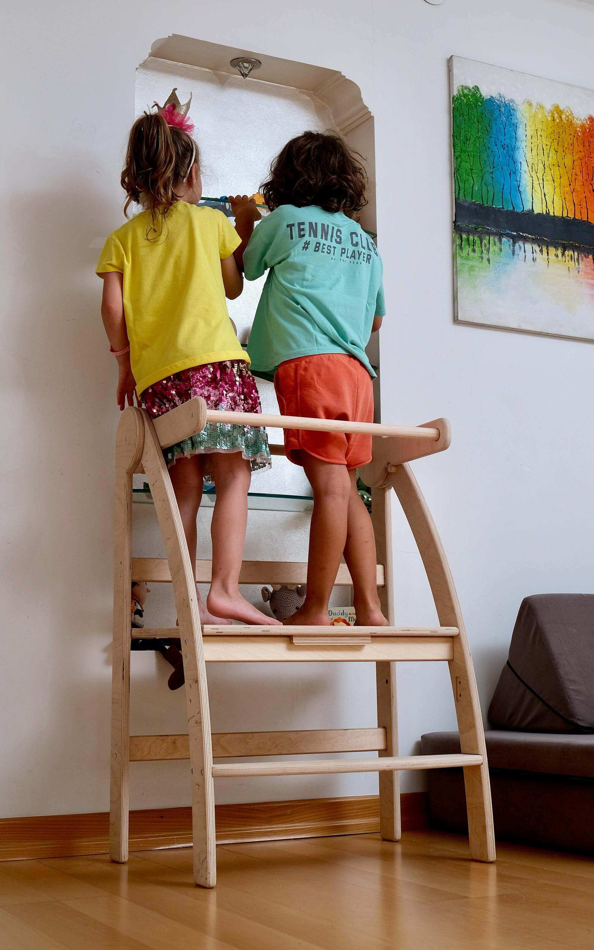 Two young children stand on a Kidodido Montessori Growth-Boosting Kitchen Tower for Kids with an adjustable and foldable design. They face a sink with a mirror, washing their hands. The child on the left wears a yellow shirt and colorful skirt, while the child on the right wears a green shirt and orange shorts. A vibrant painting hangs on the white wall behind them.