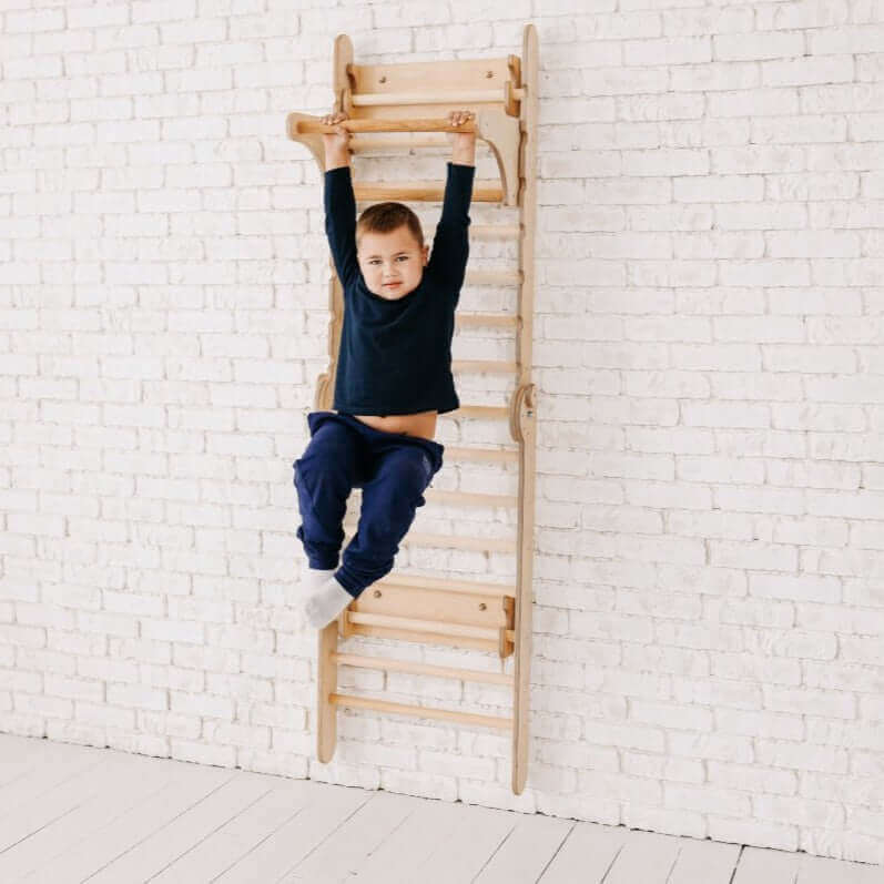 A young child in a dark blue sweater and matching pants hangs by both hands from a Goodevas Montessori Climbers Gym Addition to the Climbing Ladders, which is mounted on a white brick wall. The child’s legs are bent at the knees, with feet off the ground. The room has a light, clean, minimalist aesthetic, making it perfect for indoor entertainment.